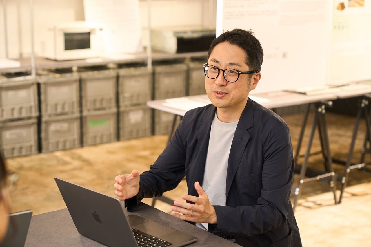 黒いジャケットと白いTシャツを着た男性がパソコンを使いながら会話をしている様子、木製の床と背景に整理された棚と展示がある