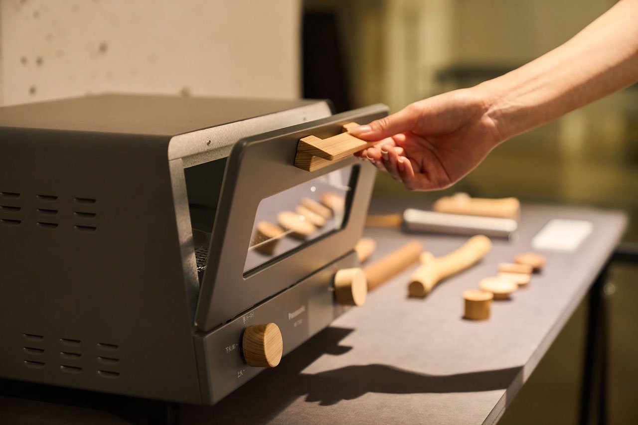 木製のトースターの取手をつかむ手、トースター前の作業台に木製の調理道具と紙袋、背景に壁と照明あり