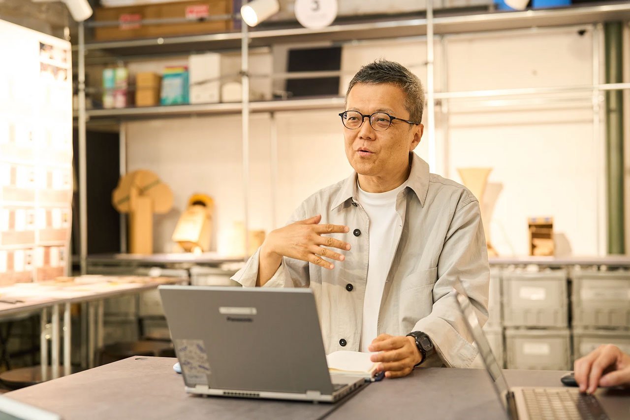 メガネをかけた男性がパソコンを使いながら手を胸の前にかざしながら話している、背景に棚と箱、窓から自然光の明るい室内