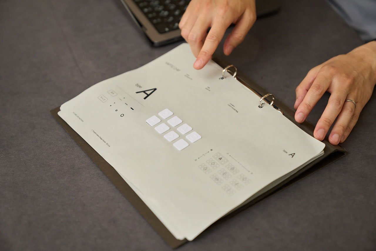 手でページをめくる、バインダーに挟まれた書類、上に白いボタン、背後にパソコン、濃い灰色のテーブル