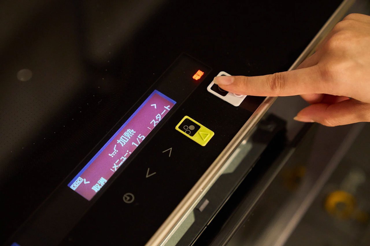 人差し指が操作パネルの白いボタンに触れる、パネルに液晶画面と操作ボタン、背景に黒い表面あり