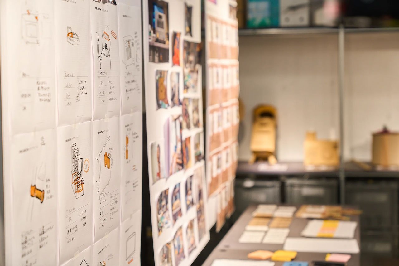 設計図やイラストが貼られた壁面と、それに伴う見本や資料の山