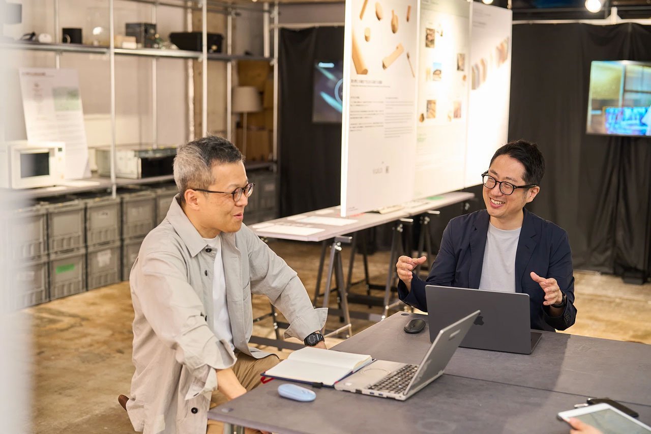 会議室のテーブルに座る二人の男性、ノートパソコンと資料を使用、背景には展示パネルとテレビスクリーン