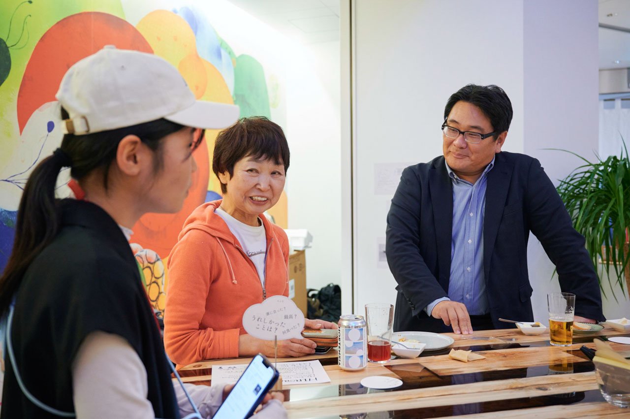 カウンターテーブルに集まる数人、オレンジのパーカーの女性と紺のジャケットの男性。背景に虹色の壁画。テーブル上に飲み物とメモ。  