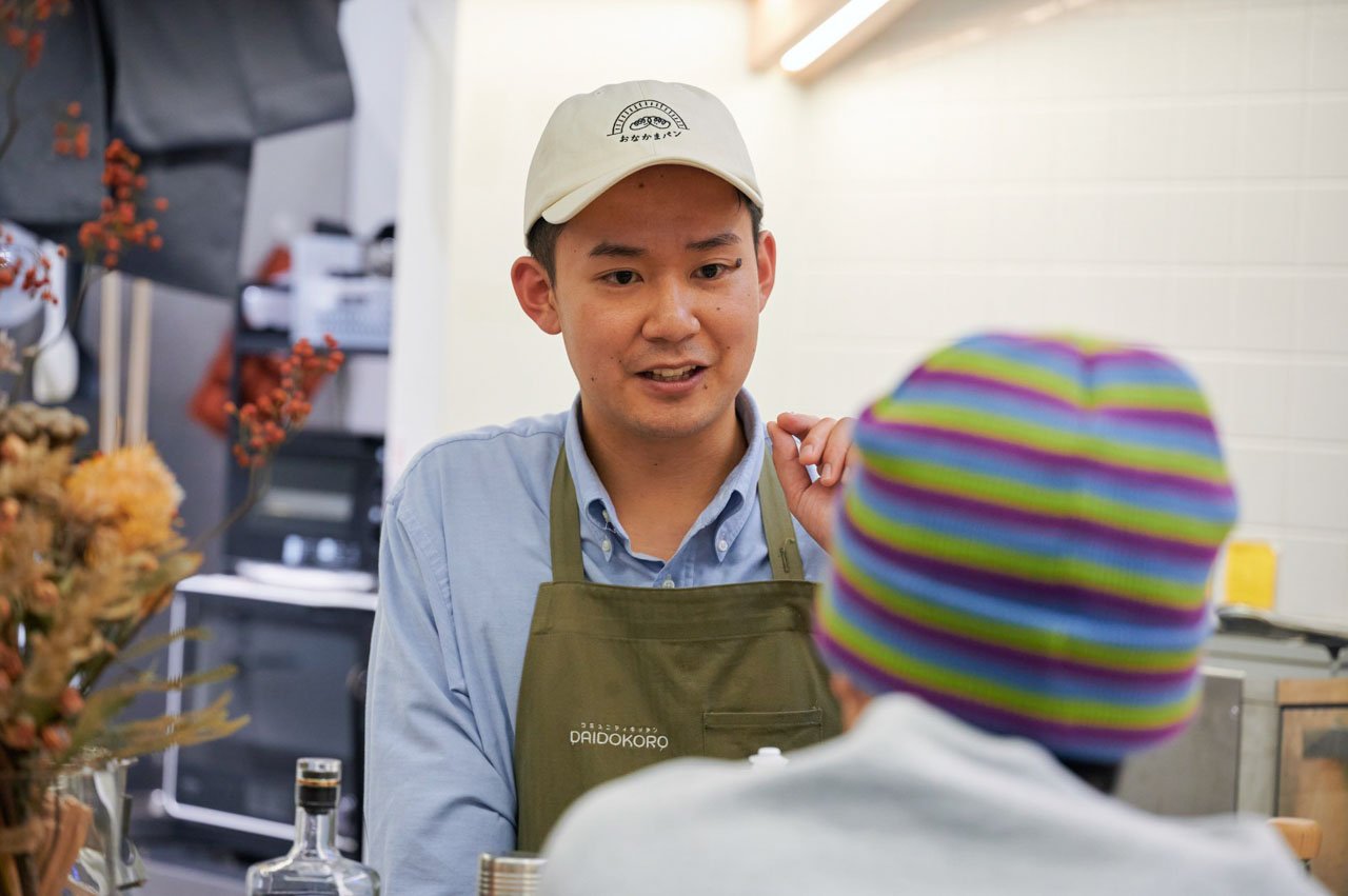 帽子をかぶった男性がカウンターでお客様と会話をしている。背景に調理器具など。