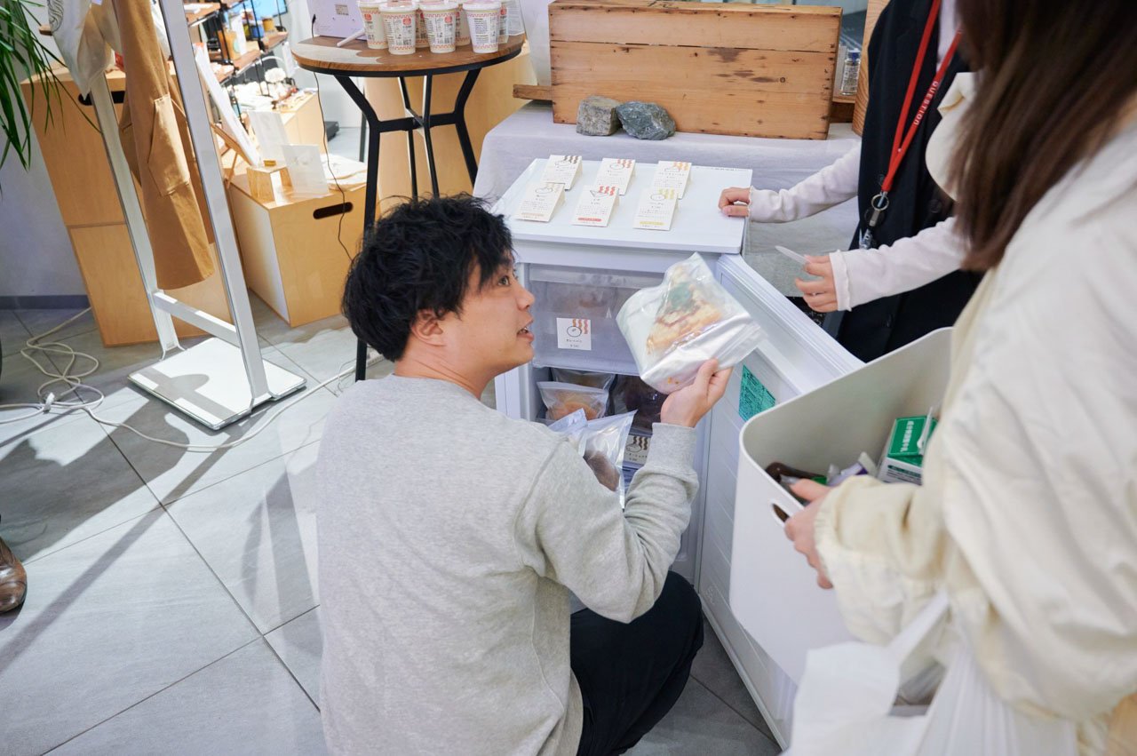 男性、灰色のセーターを着てしゃがみながら冷蔵庫から商品を取り出す。背景に商品棚。