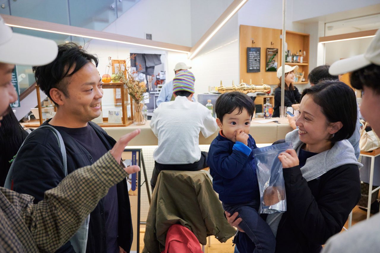 カフェのカウンター席に並ぶ人々。白いキャップ、グレーのパーカー、紺色のジャケットなど様々な服装。幼い子供を抱く人物も。