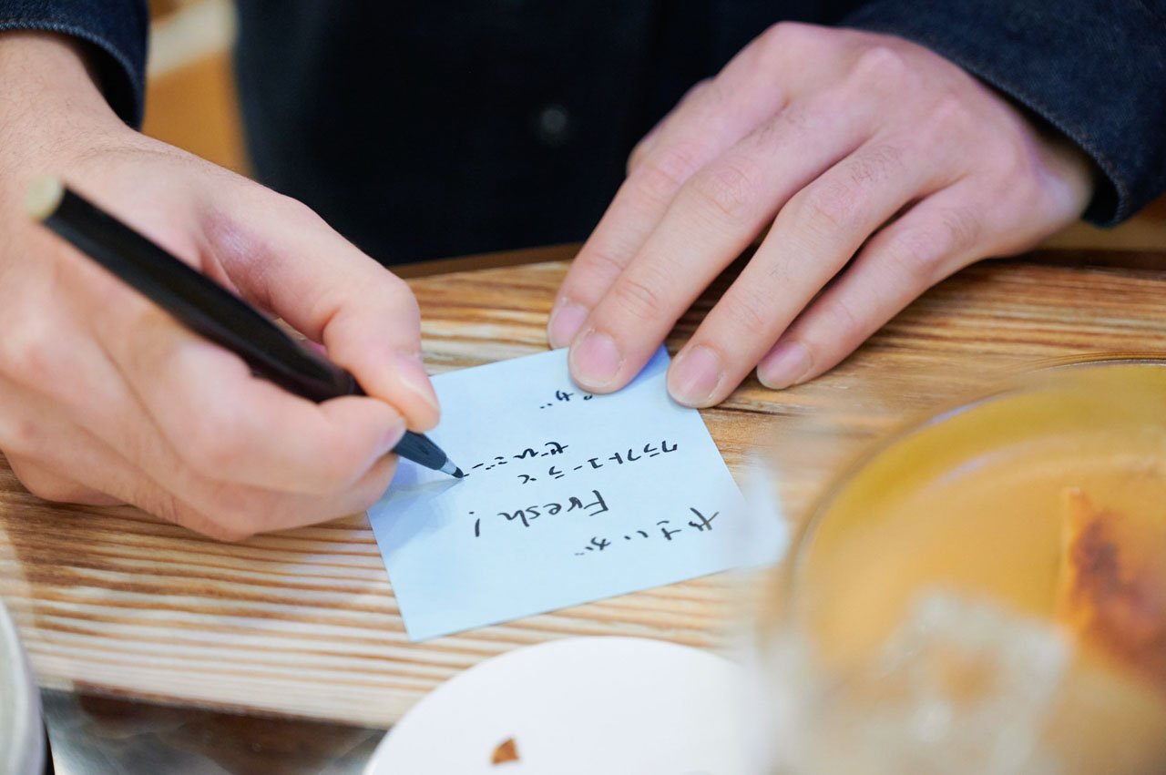 手書きのメモを取る人の手、木製のテーブル上に紙とペン、飲み物のグラスあり。