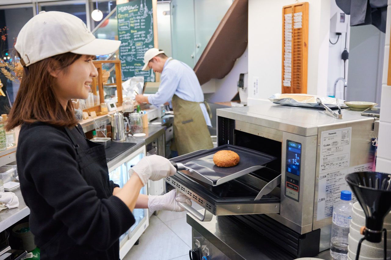 女性、白い帽子と黒い服、手袋着用、トレーにパンを乗せている。背景に厨房と人物。