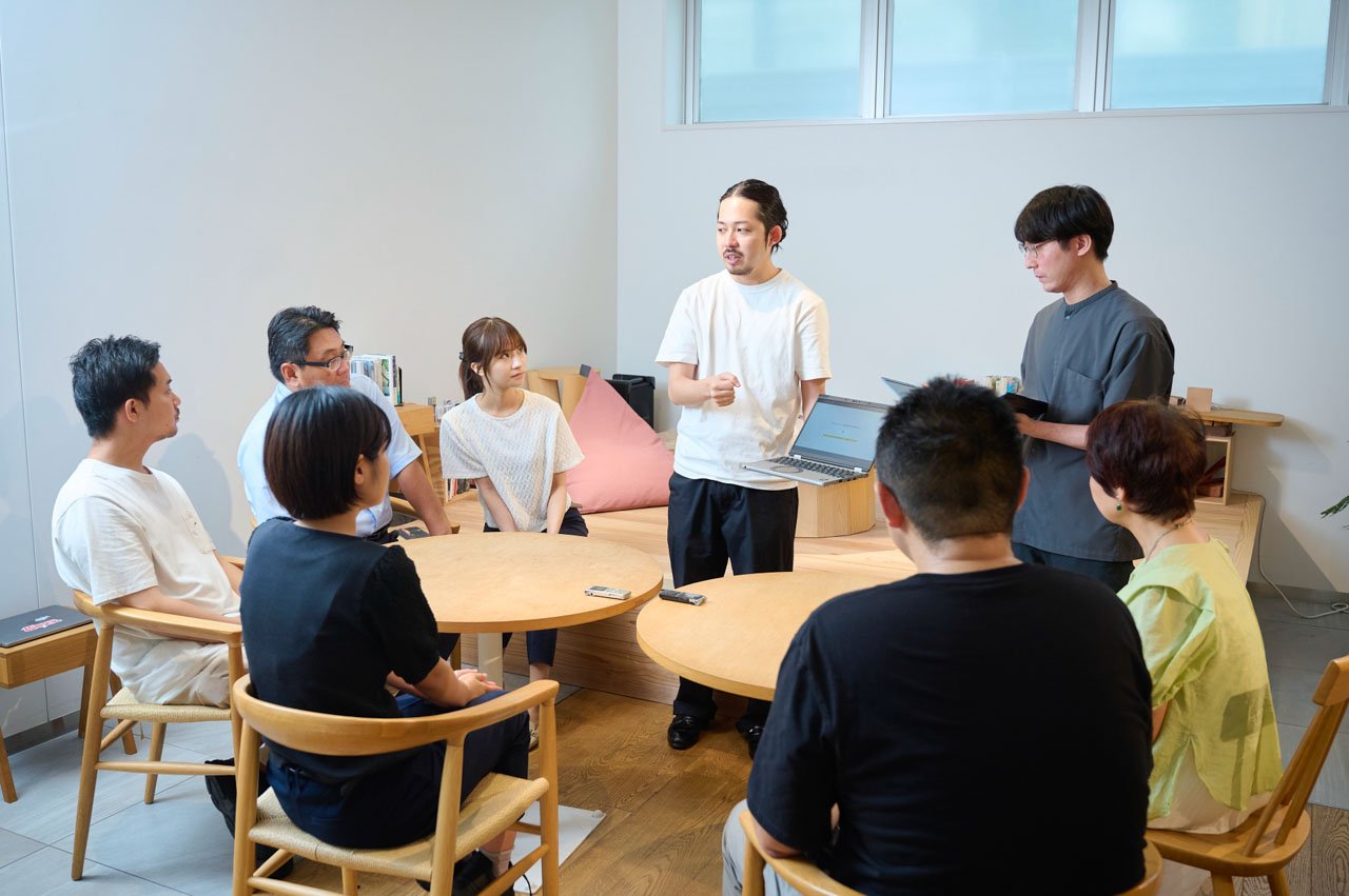 明るい室内での立ち座りの会議風景。白いTシャツ、黒いシャツ、白いニットなど様々な服装の人々。木製の椅子とテーブル。窓からの自然光。