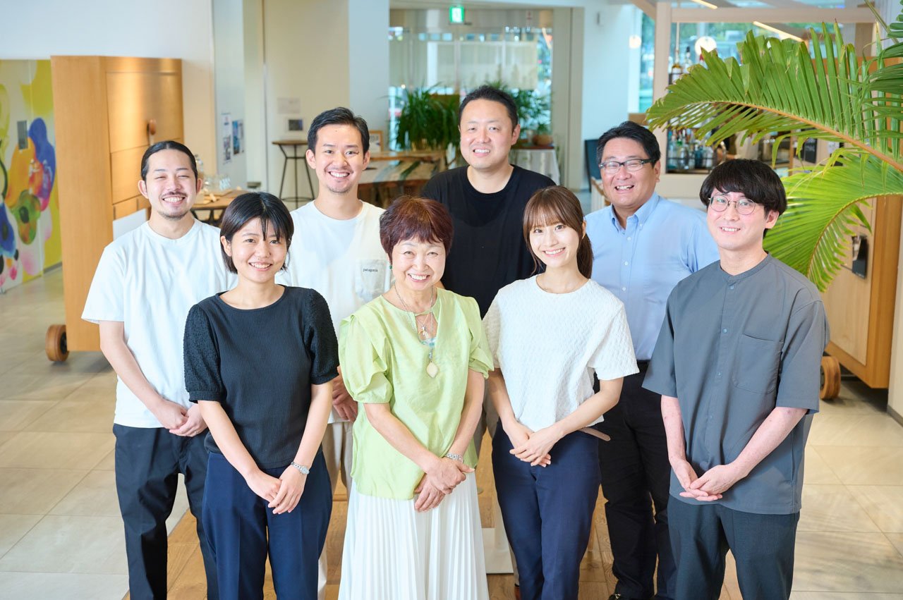 明るい空間での集合写真。白いTシャツ、黒いトップス、黄緑色の服など様々な服装の男女8名。背景に観葉植物とカラフルな壁画。