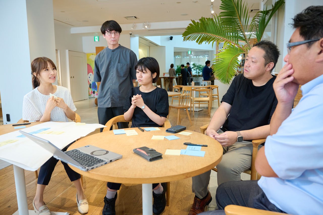 明るい室内で丸テーブルを囲む複数の人物。白いニット、黒いTシャツ、グレーのトップスなど様々な服装。テーブル上にノートPCと書類。背景に観葉植物。