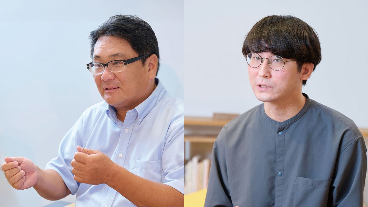 左右に人物。左に会議で話す男性の上半身、白いシャツとメガネを着用し、手振りをしている。右に灰色のシャツを着た男性、座っている。背景に白い壁と本棚。