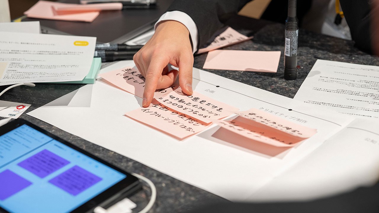 スーツを着た参加者の手元写真。ワークシートの上に意見が書かれた付箋を並べている。