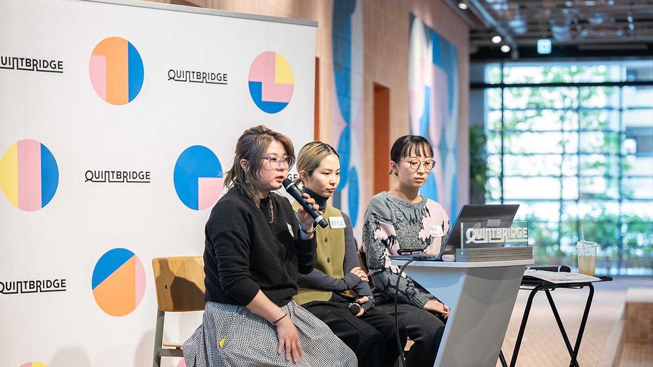 マイクを持って話す登壇者と、その隣に座る2人の登壇者を横側から撮影している。