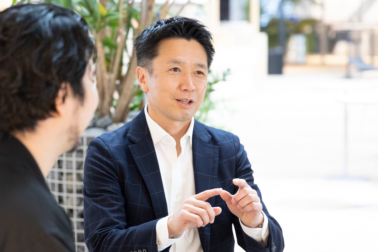 ネイビーのジャケットに白シャツの男性と黒服の別人物、屋内の観葉植物と明るい背景、顔がぼかされ手で指を示す構図
