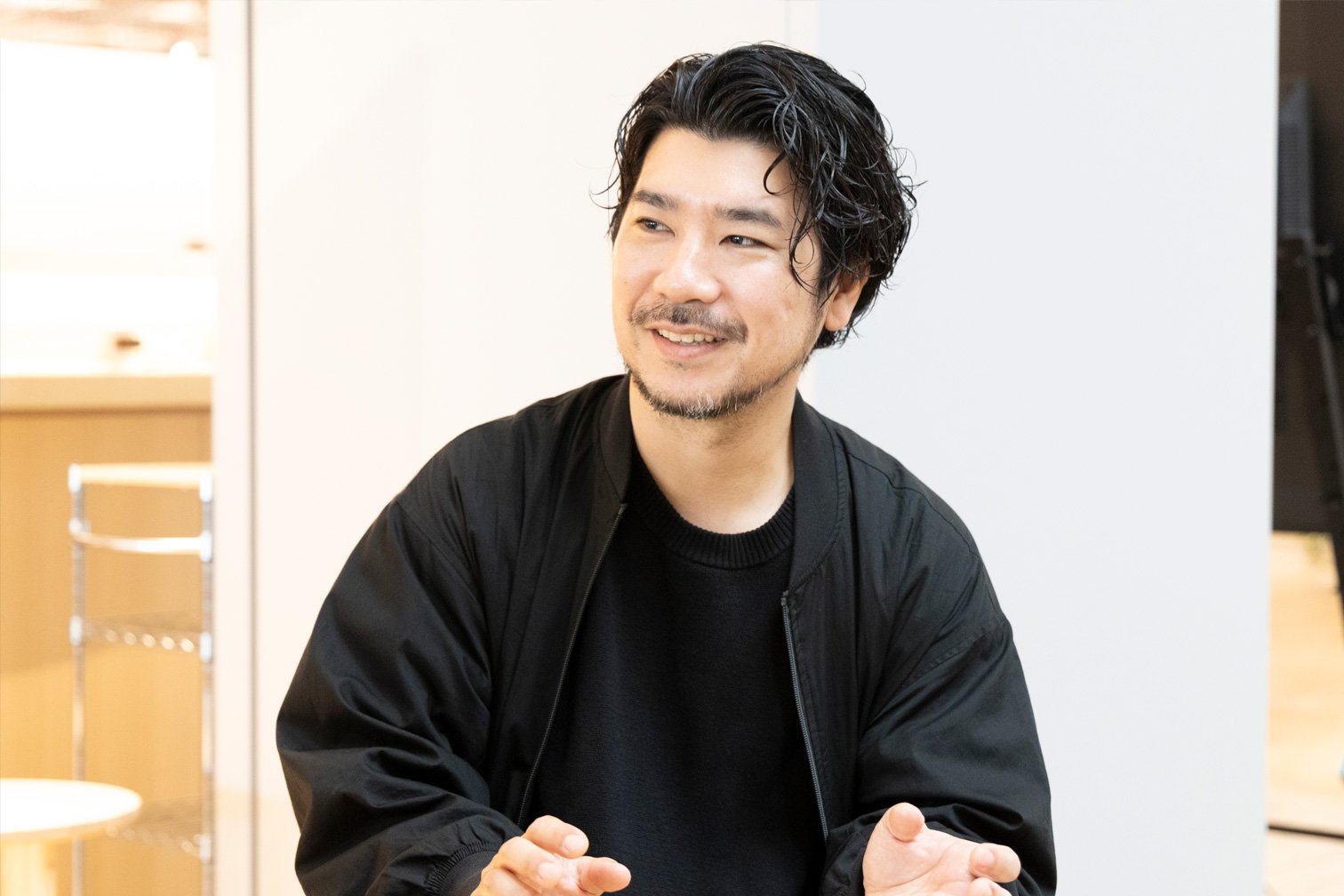 黒いブルゾンと黒シャツを着た座ってる人物、両手を前に出して話している