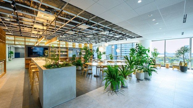 大窓と街並みの眺望を持つオープンオフィスラウンジ、格子状露出天井、木製カウンターとスツール、丸テーブルと鉢植え、タイル床