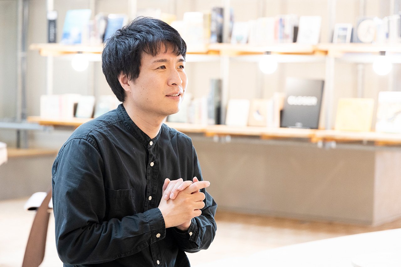 黒いバンドカラーシャツの人物、両手を組み座る姿、背後の木製棚と書籍、吊り下げ照明、木目床の室内