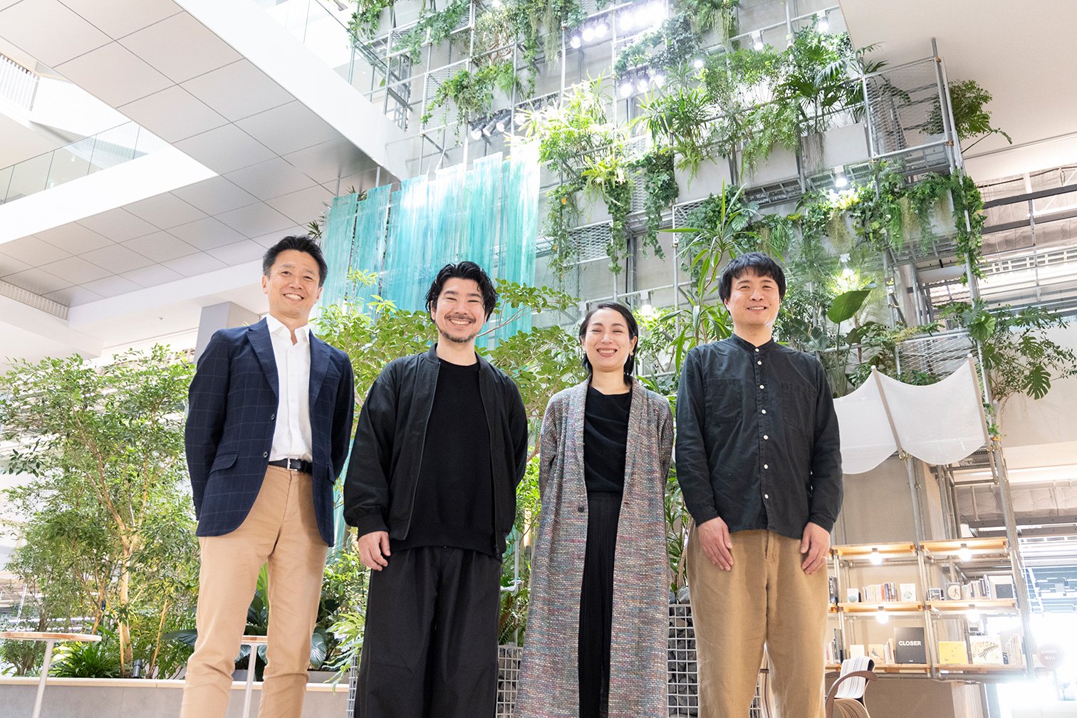 四人の人物が並んで立っている、左からネイビージャケットと白シャツ、黒ブルゾン、マルチカラーロングコートと黒ワンピース、黒ボタンシャツ、垂直緑化と水色垂れ布のアトリウム