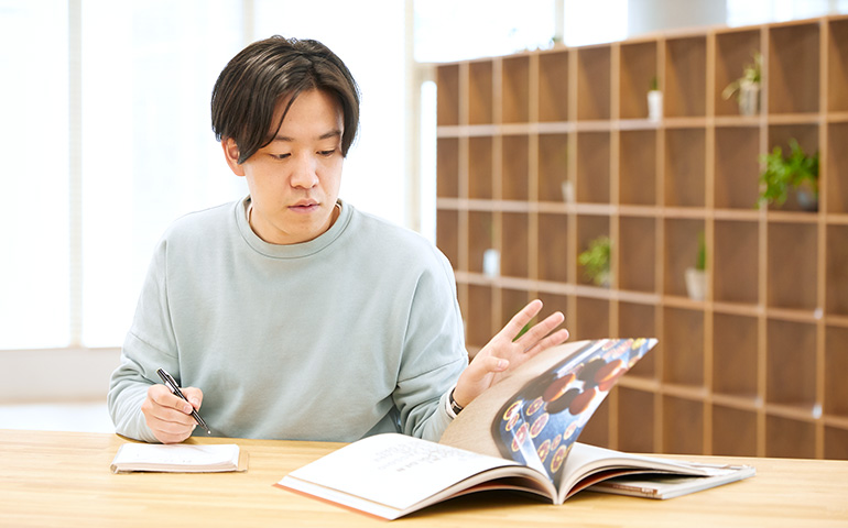 写真:テーブルで雑誌を見ながらスケッチをする飯田功太郎