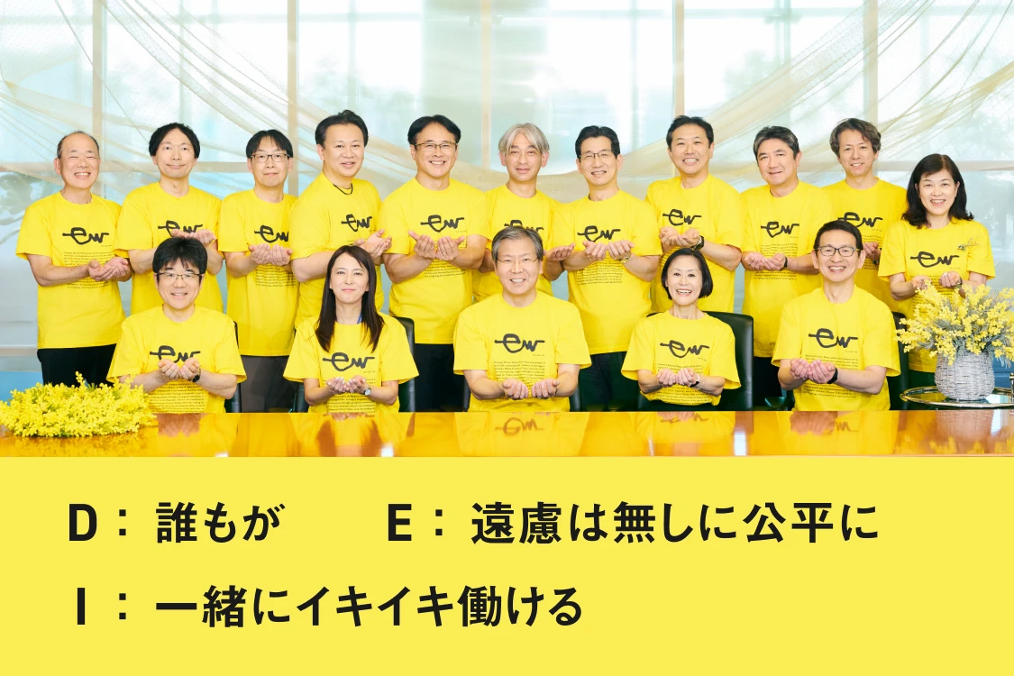写真：ミモザの花と、黄色いTシャツを着たパナソニックエレクトリックワークス役員の集合写真 D：誰もが E:遠慮は無しに公平に I:一緒にイキイキ働ける