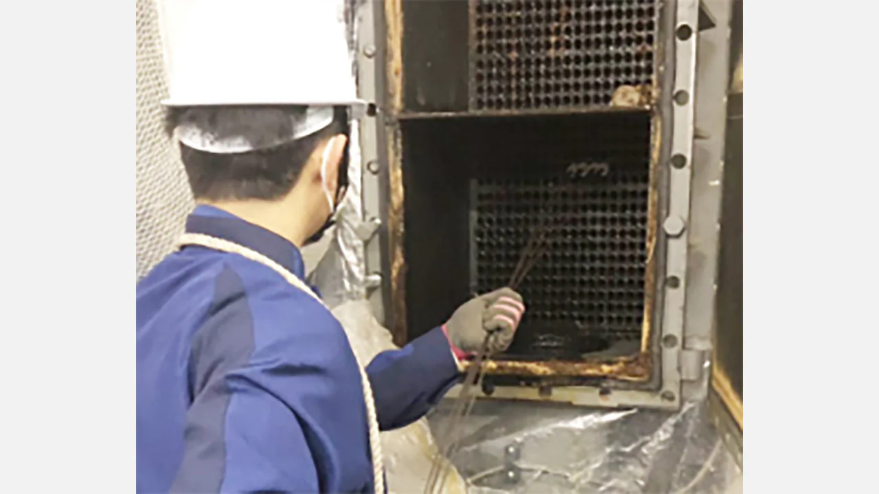 伝熱管ブラシ洗浄の作業写真。ヘルメットとマスクを着用した作業員が、開口した機械内部の管板に並ぶ伝熱管へ洗浄ブラシを差し込み、内部の汚れを落としている。