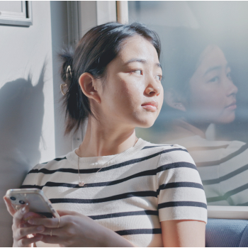 窓から差し込む光を浴びながら、スマートフォンを持って窓の外を見つめる女性