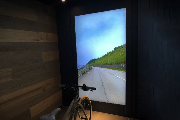 エア クリエイションの画像。床に自転車が置かれており、自転車の前の大きなスクリーンに緑が見える道路が映し出されている。