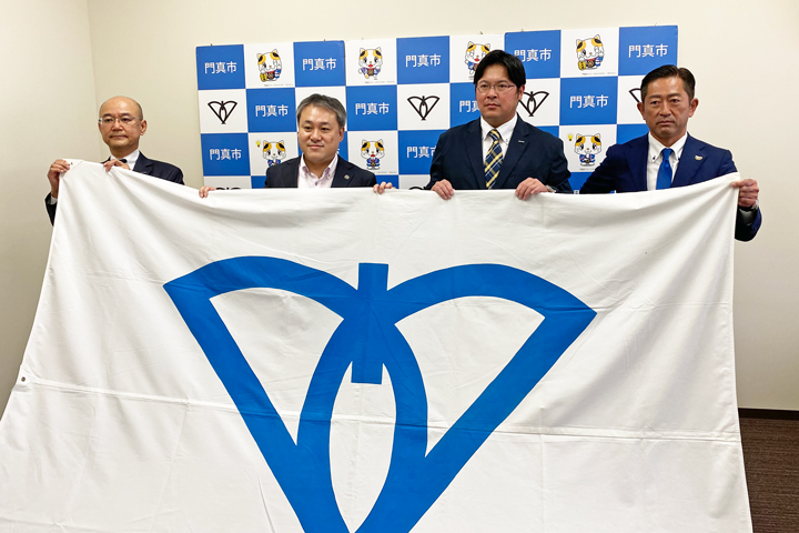 画像：（左から）市旗を掲げる羽田野部長、宮本市長、田中監督、久保田所長