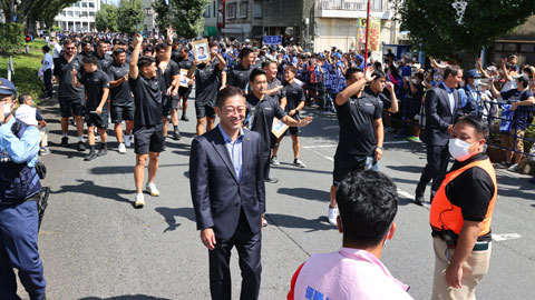 画像：パレードに参加できなかったメンバーの写真を持ちながらファンに手を振るメンバーたち。